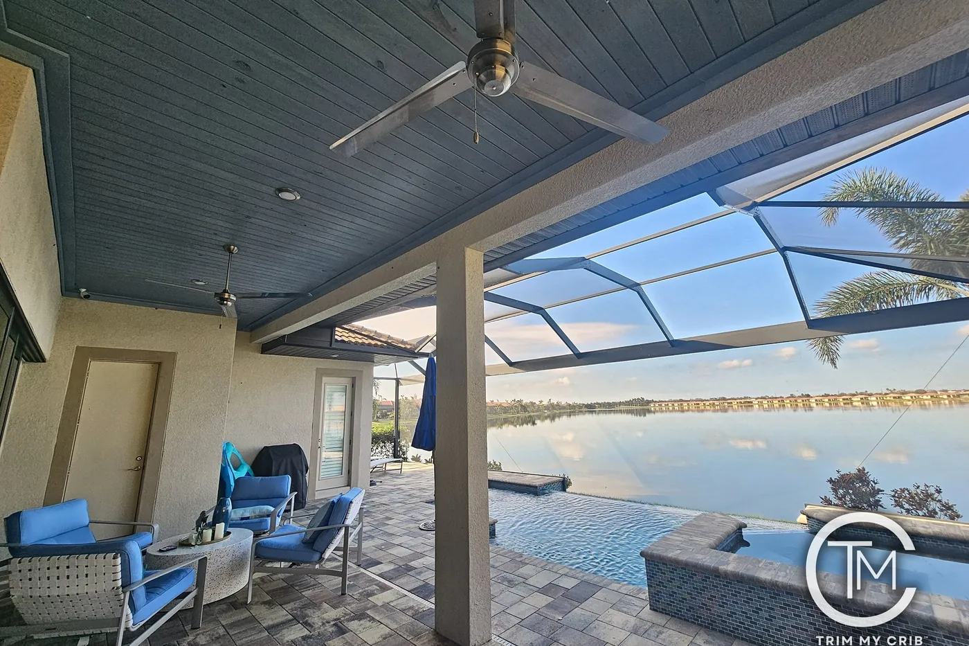 Lanai with a painted gray plank ceiling, ceiling fan and blue chairs overlooking a lake through the pool cage